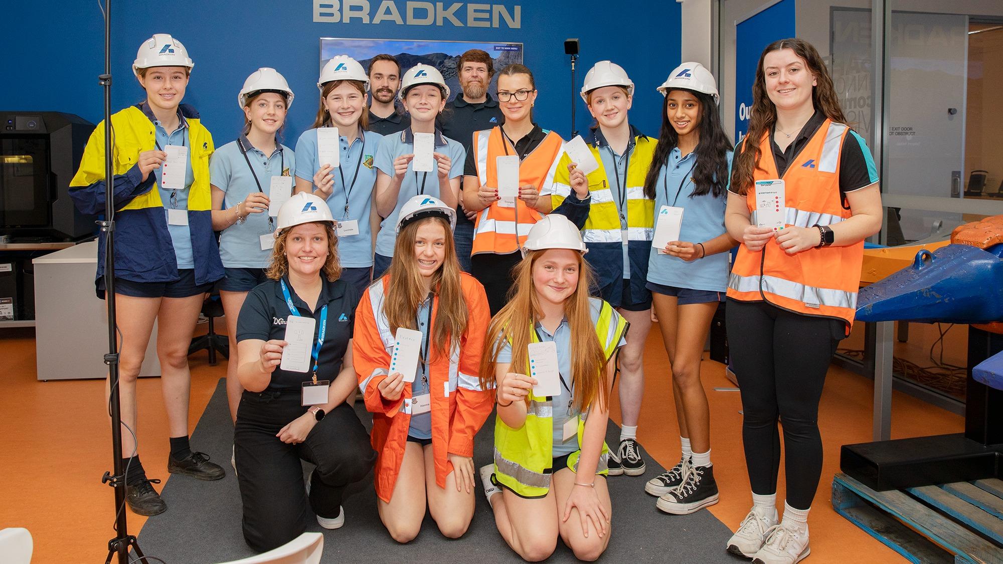 A group of school girls wearing Bradken branded hard hats and hi-viz vests over their uniforms stand with Bradken team members wearing Bradken branded polo shirts. All hold small pieces of paper displayed to the camera.