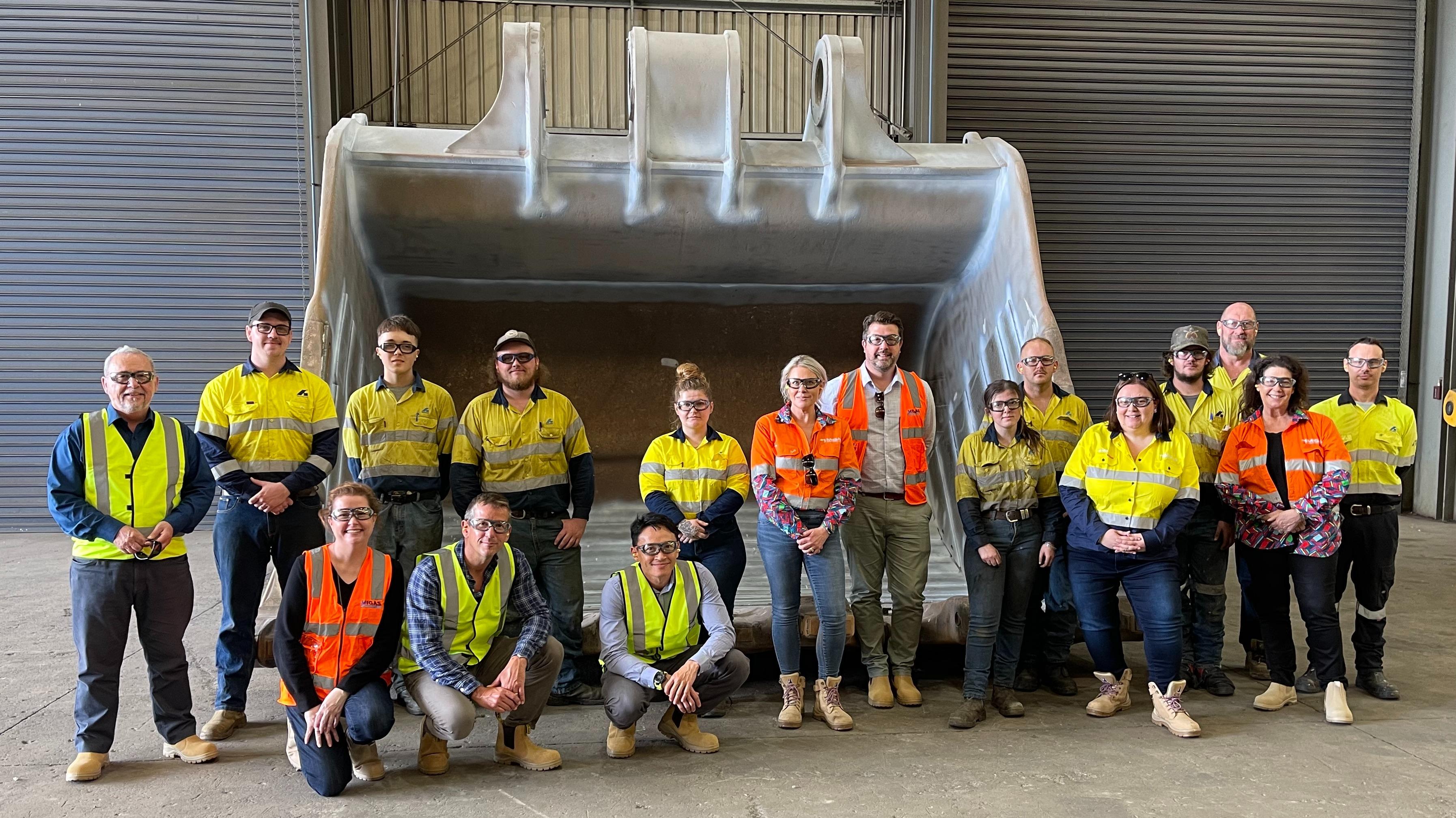 A group of 17 men and women wearing hi-vis clothing stand in front of an excavator bucket inside a workshop.