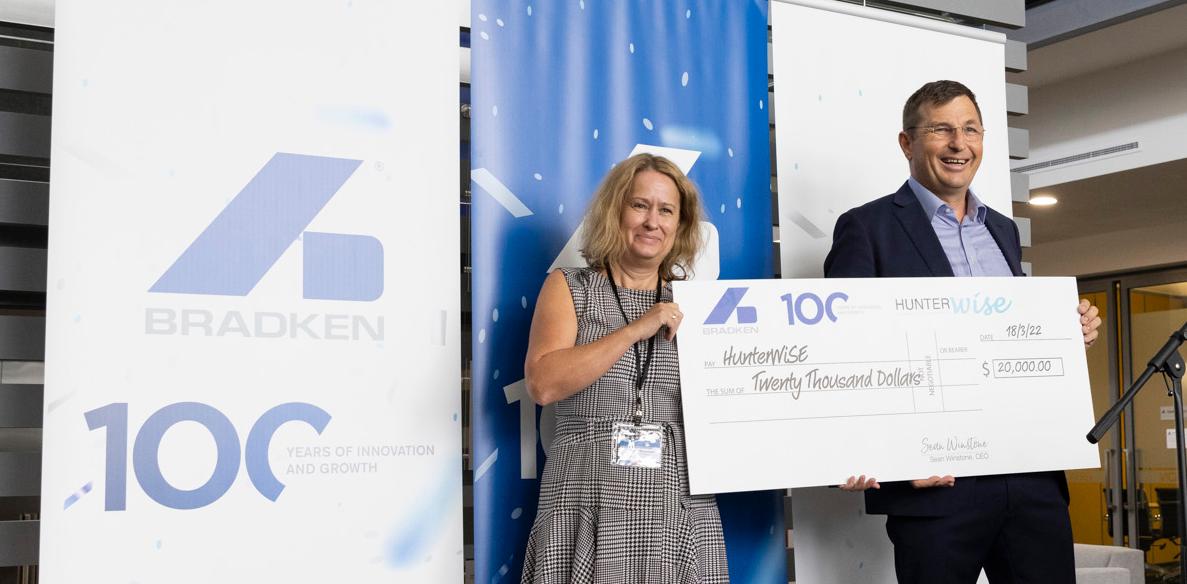 A woman wearing a black and white checked dress stands next to a taller man in a suit. Both are holding the same novelty cheque and are standing in front of blue and white banners with the Bradken logo on them.
