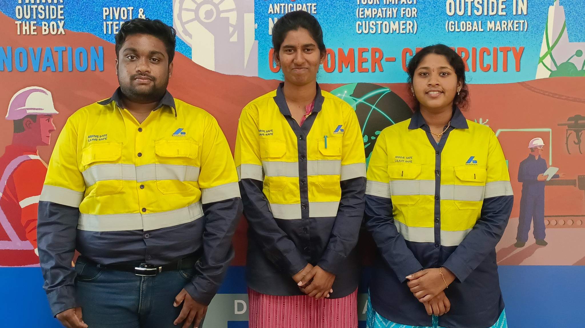 A man and two women stand in line shoulder to shoulder, wearing yellow hi-vis shirts with the Bradken logo on them, against a colourful banner backdrop.