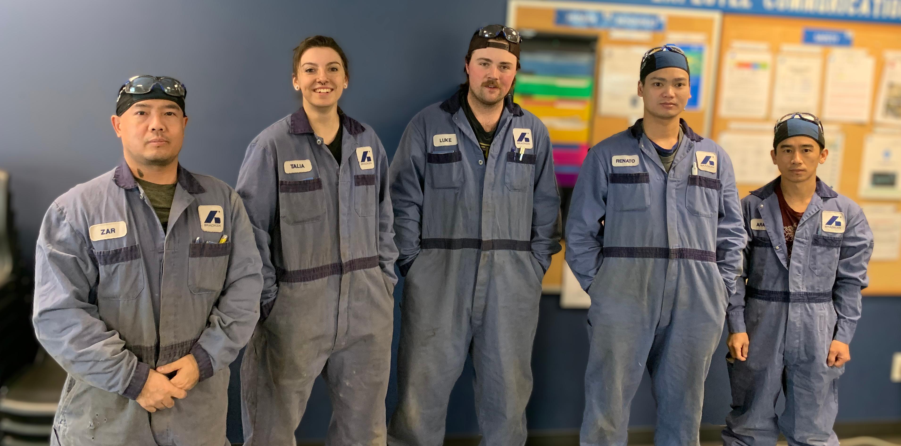 Five workers, four men and one woman, stand in a row. All are wearing blue coveralls with the Bradken logo on them.