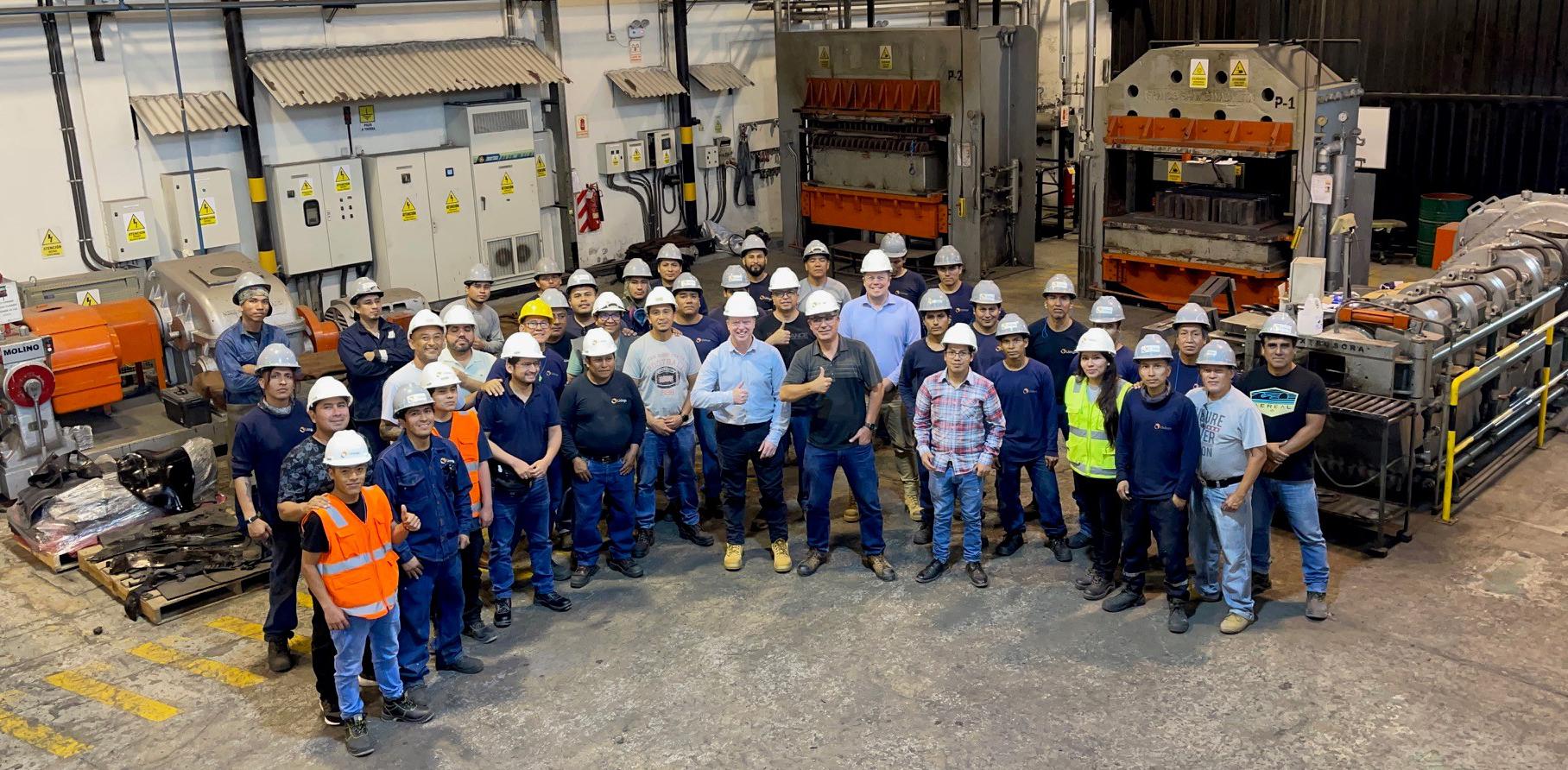 A team of factory employees stands together in a workshop. They are wearing white hardhats and some are giving the thumbs up hand gesture.