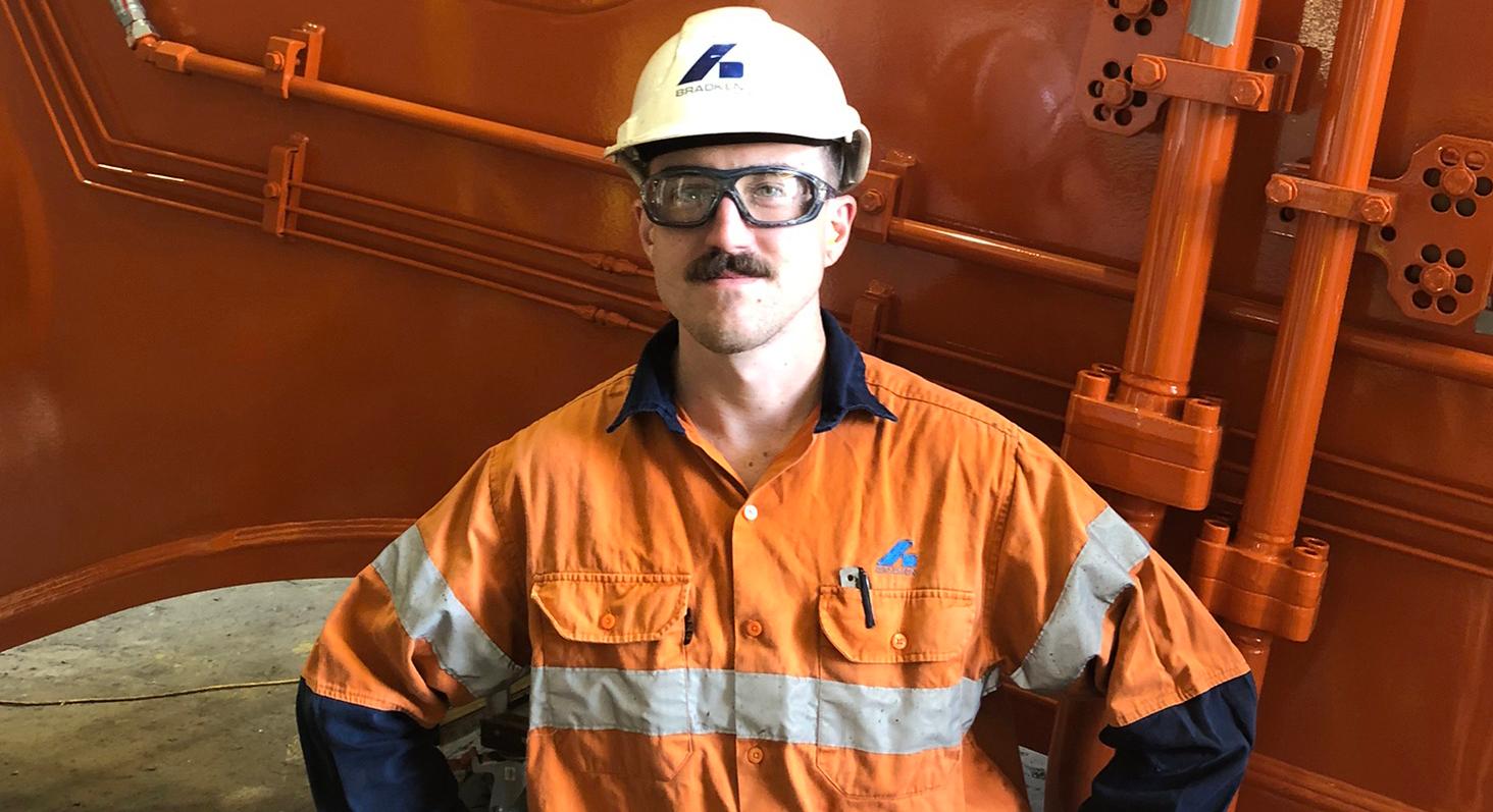 man in orange hi-visibility work shirt, safety glasses and a white hard hat with the Bradken logo stands facing camera with his hands on his hips