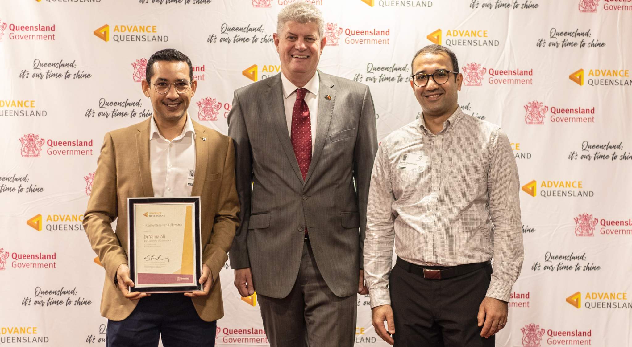 Three people, all men, stand in front of a media wall - the man on the left holds a framed certificate.